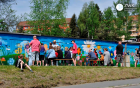 Eine altersgemischte Gruppe Menschen steht an einer langen Mauer und bemalt sie mit einem Motiv aus Blumen, Wiesen und Insekten. 