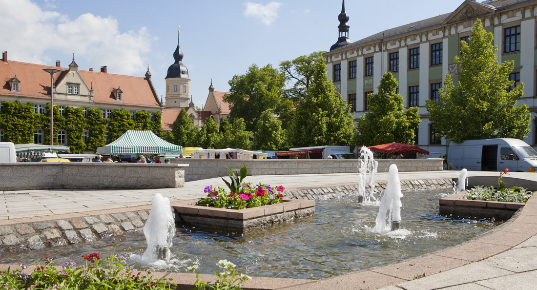 Ein öffentlicher Platz mit Wasserspiel, Blumen und einem Markt im Hintergrund.