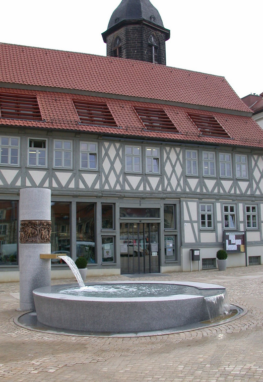 Ein grauer, flacher Brunnen vor einem großen Fachwerkhaus mit rotem Dach.