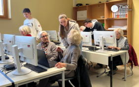 Einige lebenältere Menschen sitzen an Tischen vor Computern. Zwischen den Tischen stehen drei Jugendliche.