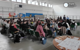 Viele Schülerinnen und Schüler sitzen in einer Sporthalle. Auf einem Tisch liegen Zeichnungen und Pläne.