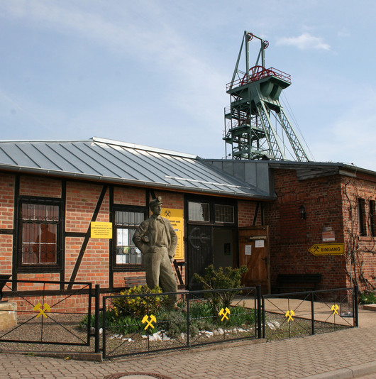 Ein Fachwerkhaus mit roten Ziegeln. Im Vorgarten die Bronzefigur eines Bergbauarbeiters. Im Hintergrund der Röhrigschacht.