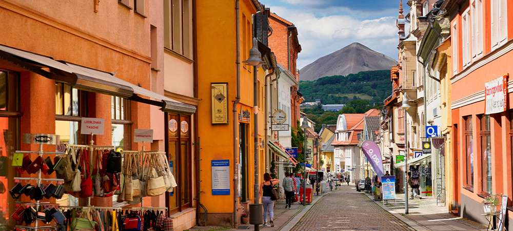 Eine Straße mit Kopfsteinpflaster und kleinen Läden in Wohnhäusern. Im Hintergrund ist ein Berg zu sehen. Die Sonne scheint und wirft Schatten.