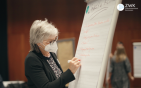 Eine Frau mit kurzen weißen Haaren steht einer Flipchart und notiert etwas 