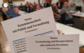 Im Hintergrund sitzen Menschen an Tischen. Im Vordergrund sind zwei Zettel zu sehen auf denen die Themen der nächsten Veranstaltung stehen.