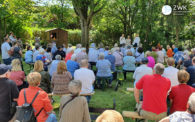 160 Menschen sitzen im Freien auf Bänken und nehmen an einer Versammlung teil.