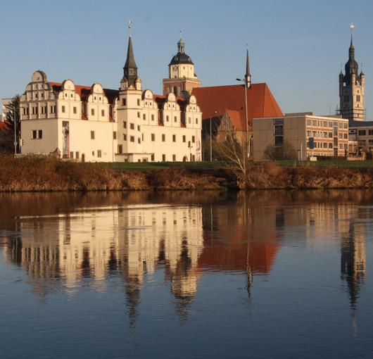 Ein Schloss spiegelt sich in der Dämmerung in einem See. Im Hintergrund sind Gebäude der Stadt zu sehen.
