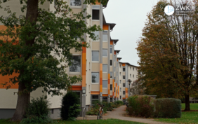 Ein Wohngebäude mit vier Etagen und orangener Fassade. Die Umgebung ist mit Bäumen und Wiesen begrünt.