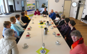 Zwölf ältere Damen und zwei Herren sitzen bei Kaffee und Erbeerkuchen an einem Tisch. Im Hintergrund ein Roll-Up der ZWK.