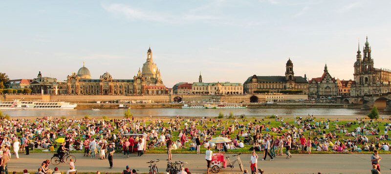 Ein Flussufer, viele Menschen die gemeinsam feiern und im Hintergrund barocke Gebäude.
