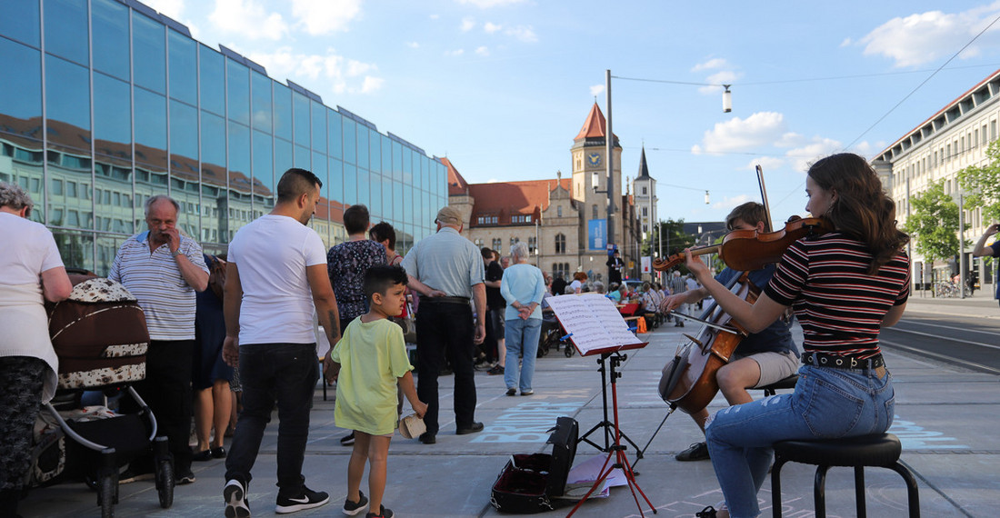 Rechts sitzen zwei junge Menschen und spielt klassische Instrumente, links gehen Menschen an einer verglasten Fassade vorbei.