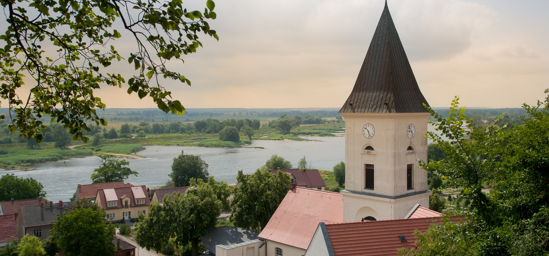 Ein schlichter Kirchturm mit dunklem Dach in einem kleinen Dorf. Im Hintergrund ein Fluss und grüne Landschaft.