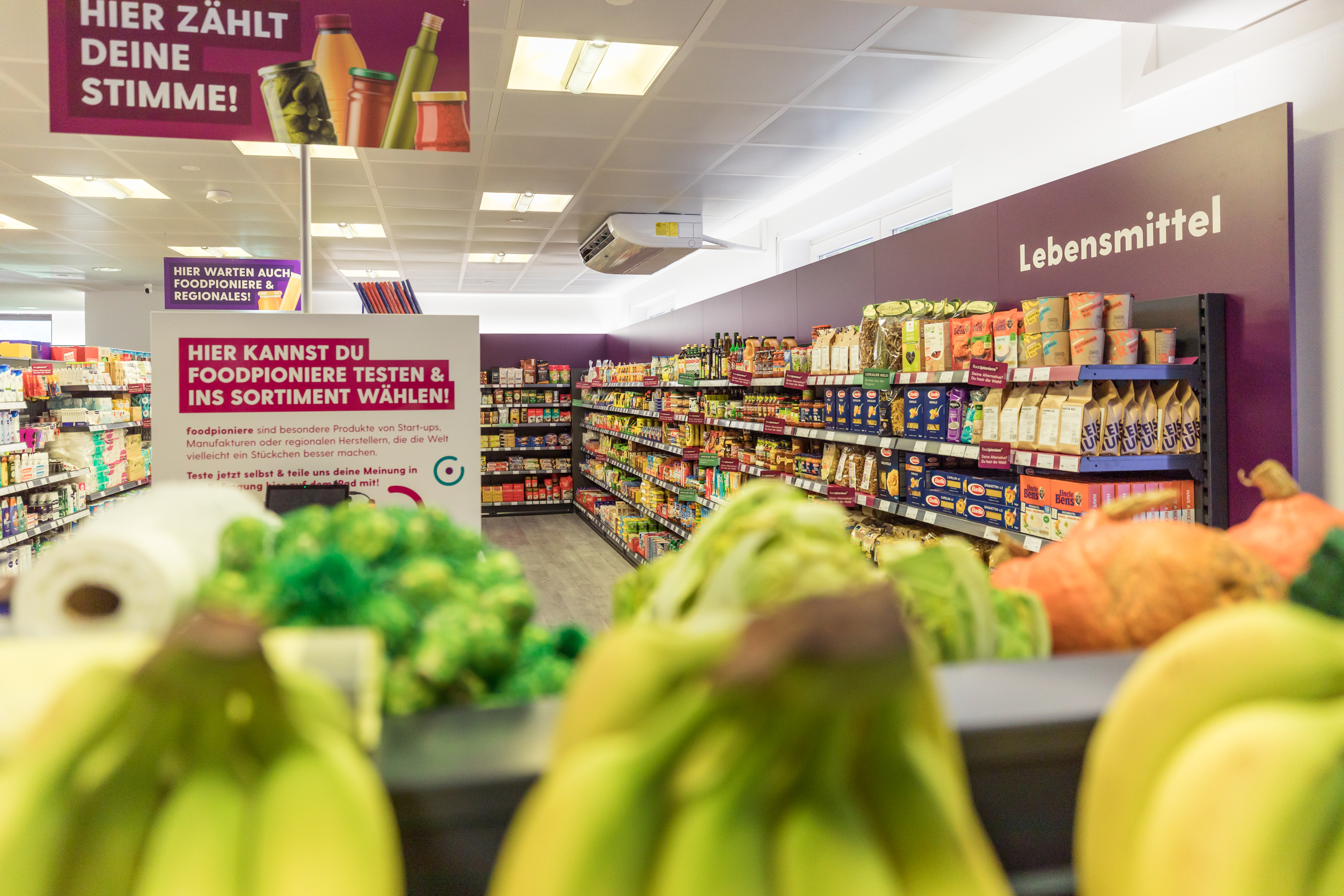 Blick in einen Supermarkt: Bananen im Vordergrund und gefüllte regale sowie ein digitaler Counter mit dem Hinweis "Hier zählt deine Stimme - Hier kannst du Foodpioniere testen und ins Sortiment wählen"