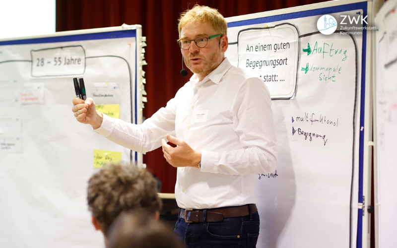 Eine Person in weißem Hemd steht vor Flipcharts und hält dabei zwei Stifte in der Hand.