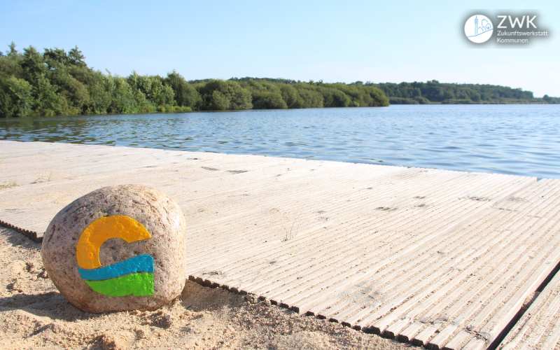 Ein Stein mit dem Logo von Geestland liegt auf einem Steg, dahinter ein See und Bäume