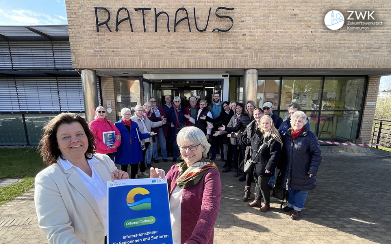 Im Vordergrund zwei Frauen, die ein Plakat zur Seniörenbörse in die Kamera halten. Im Hintergrund eine Gruppe Teilnehmende.