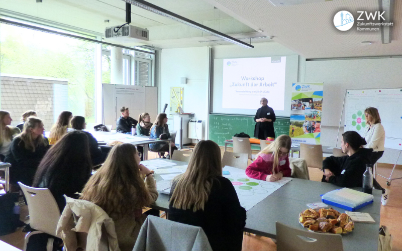 Mehr als ein Dutzend Studierende sitzen an Tischen und hören einer mittelalten Frau zu, die vor einer Präsentation steht. Im Raum ist auch ein Roll-Up der ZWK zu sehen.