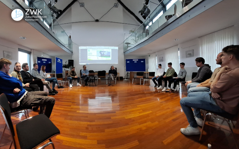 Ein Gruppe junger Menschen sitzt in einem großen Saal mit sehr hohen Decken im Stuhlkreis. Im Hintergrund eine Leinwand mit einer Präsentation.