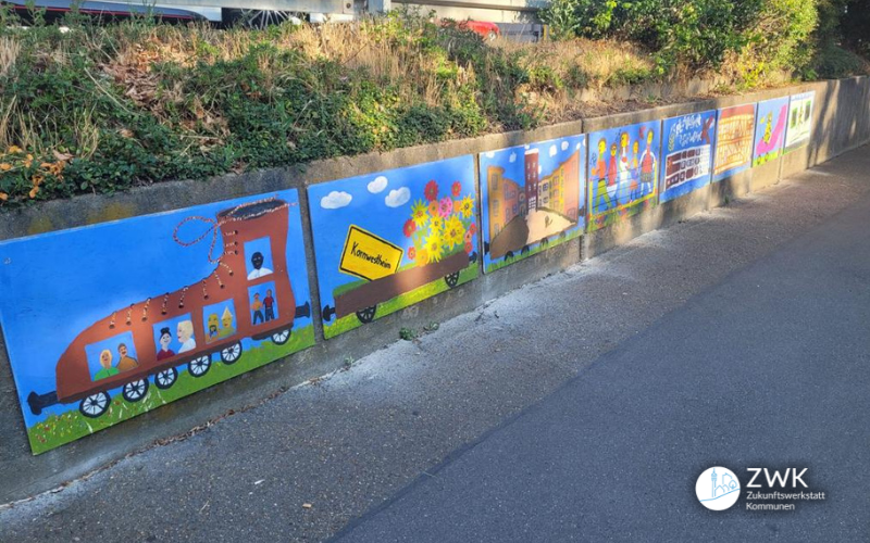 Eine Mauer mit farbigen Bildern. Ein Zug sowie blauer Himmel sind abbgebildet. Dahinter eine bepflanzte Böschung und eine Leitplanke.