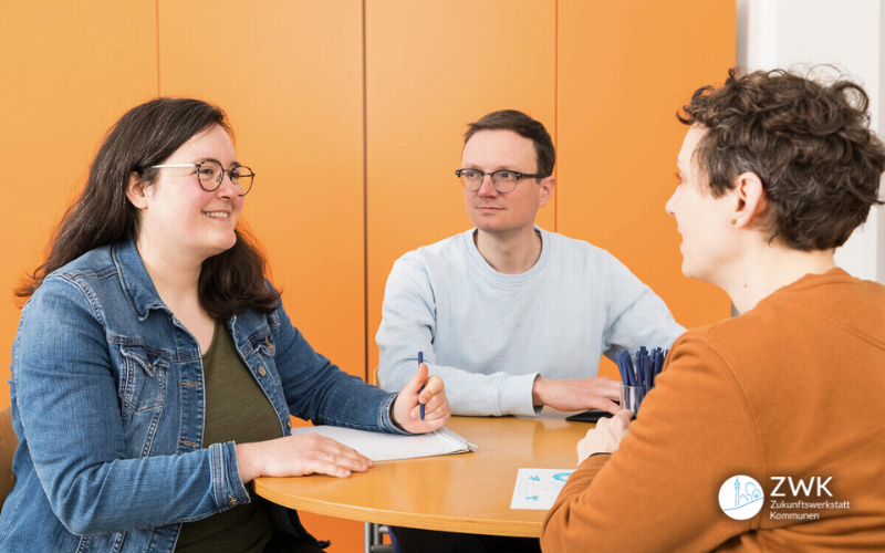 Drei junge Erwachsene sitzen um einen Tisch und diskutieren. Im Hintergrund ein orangefarbener Schrank, auf dem Tisch blaue Stifte und ein Heft.