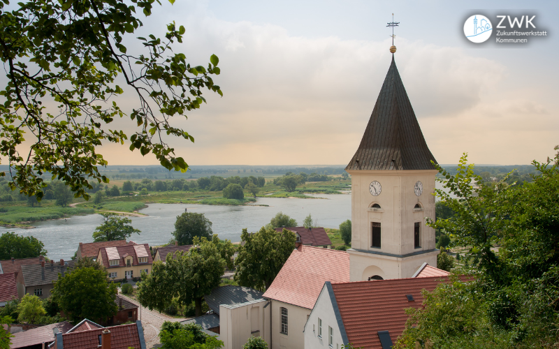 Ein schlichter Kirchturm mit dunklem Dach in einem kleinen Dorf. Im Hintergrund ein Fluss und grüne Landschaft.