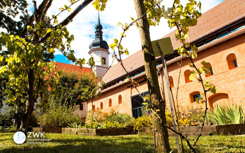 Ein Klostergebäude mit Kirchturm, Beeten und einer Weinrebe im Vordergrund.