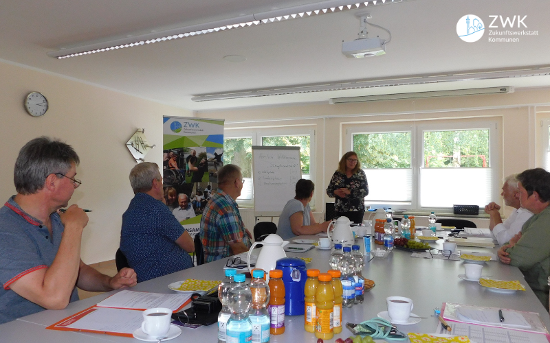 Sechs Erwachsene sitzen um einen Tisch. Eine Frau trägt etwas vor einer Flipchart vor.