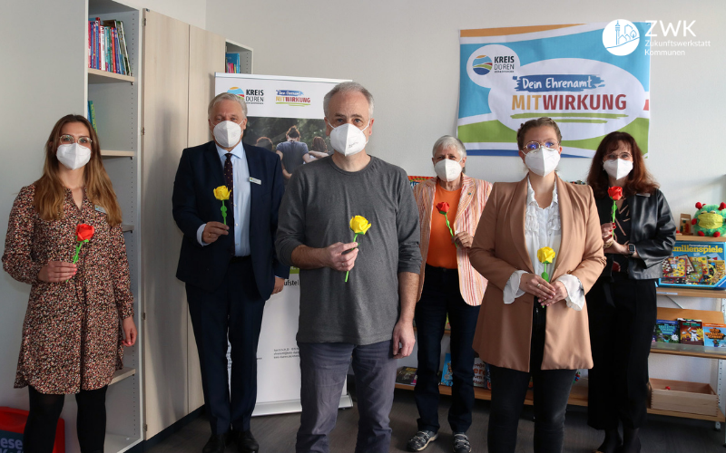 Vier Frauen und zwei Männer mit gelben Blumen in der Hand, stehen in einem Büro. Im Hintergrund ein Plakat mit der Aufschrift: Dein Ehrenamt MitWirkung