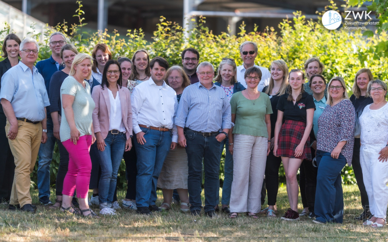 Eine Gruppe von Frauen und Männern mittleren Alters posieren für ein Foto. Sie stehen bei Sonnenschein draußen auf einer Wiese.