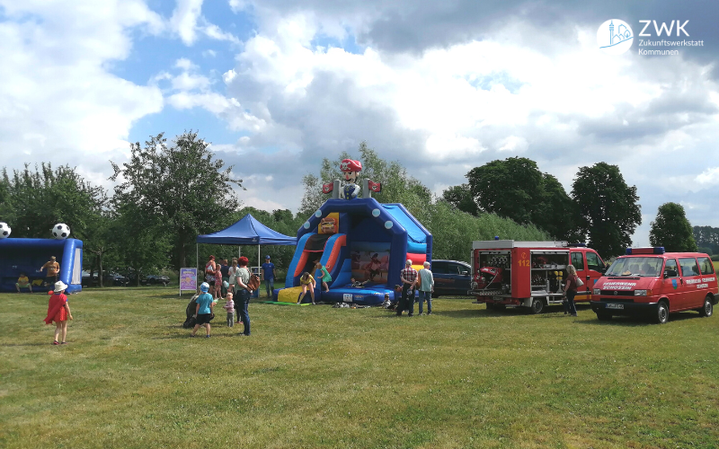 Eine große Wiese mit Kindern, einer Hüpfburg, einem Feuerwehrauto und einer Torwand.