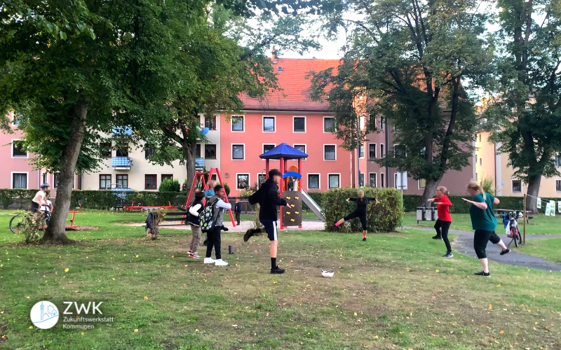 Kinder und Erwachsene, die auf einem Spielplatzgelände stehen und gemeinsam Sportübungen machen.