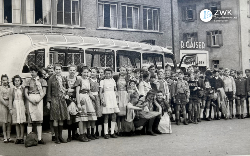 Historisches Bild in schwarz-weiß. Mehrere Dutzend Kinder stehen für ein Foto vor einem Bus. Hinter dem Bus ist ein großes Gebäude zu sehen.